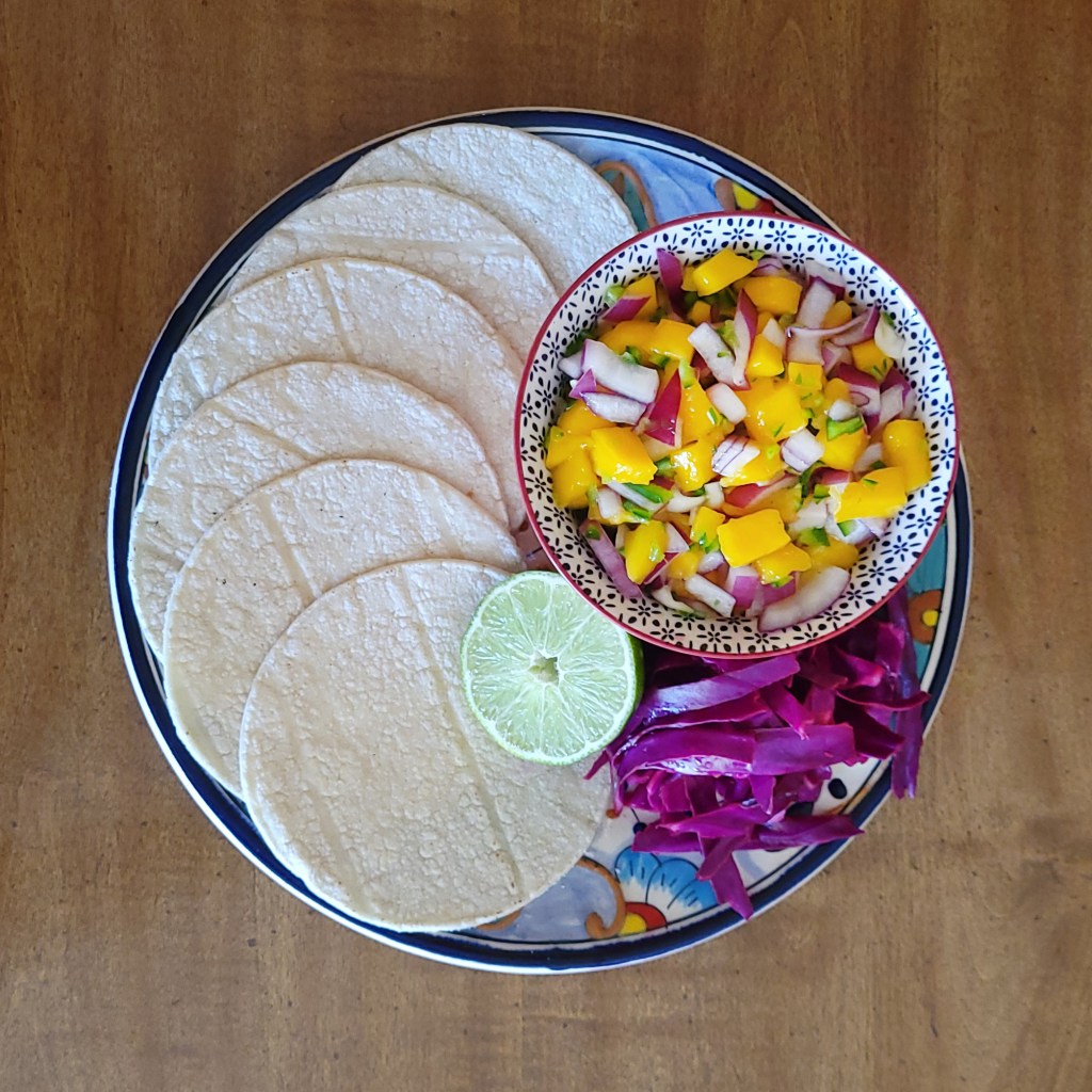 A plate displaying accompanying ingredients: corn tortillas, a bowl of mango salsa, lime half, and strips of red cabbage