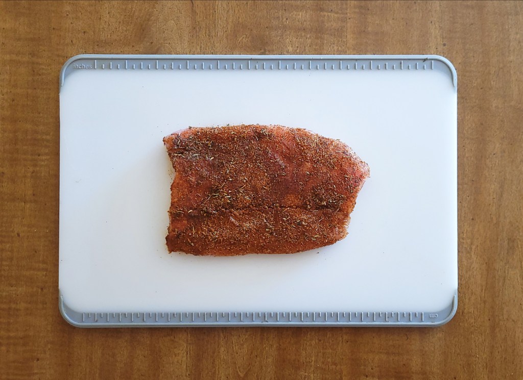 A raw corvina fillet covered in blackening seasoning sits on a while cutting board, ready for cooking.
