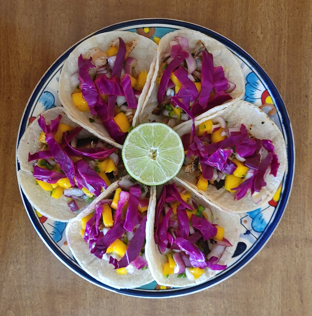 A plate of six corvina tacos fully assembled in a circle like the petals of a flower, with the lime half decoratively in the center.